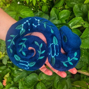 May include: A plush, coiled snake toy in a deep blue hue, adorned with embroidered green leaves and a constellation design. The soft toy is held in a hand, showcasing its detailed craftsmanship and whimsical design, perfect for a child's toy collection.