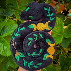 May include: A plush, black snake-shaped toy with green leaf and key embroidery. The snake features yellow crescent moon and full moon designs. The toy is made of soft, dark fabric and is designed to be cuddled.
