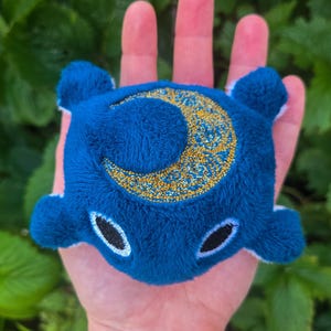May include: A plush, blue creature with a crescent moon design. The moon is gold with intricate patterns. The creature has two black oval eyes and small white accents. The plush is held in a hand against a green leafy background.