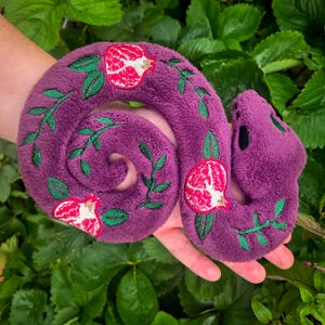 May include: A plush purple snake toy with embroidered details. The snake features red and white pomegranate designs and green leaf accents. The snake is coiled and has a rounded head with black eyes.