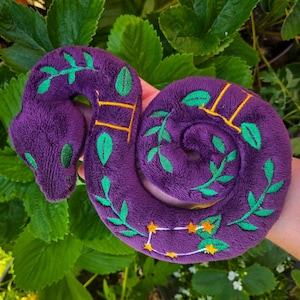 May include: A plush purple snake toy with green leaf and yellow star embroidery. The snake is coiled and has green eyes. The toy is made of soft material and is designed to be a child's toy.