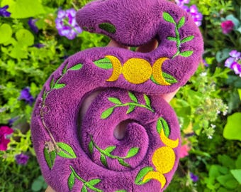 May include: Plush purple snake toy with embroidered yellow crescent moons and green leaves. The snake is coiled and has a soft, fuzzy texture. The background is a garden with green foliage and colorful flowers.