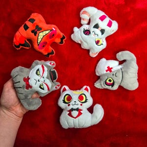 May include: Five plush cat toys in various designs, including a red and black creature, a white cat with pink accents, and gray cats with unique facial features. The toys are arranged on a red surface.