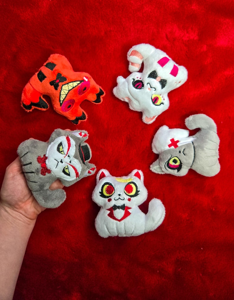 May include: Five plush cat toys in various designs, including a red and black creature, a white cat with pink accents, and gray cats with unique facial features. The toys are arranged on a red surface.