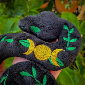 May include: A plush black snake toy featuring embroidered details. The design includes a central gold triple moon symbol flanked by vibrant green leafy vine patterns that wrap around the soft, textured fabric of the coiled serpent body.