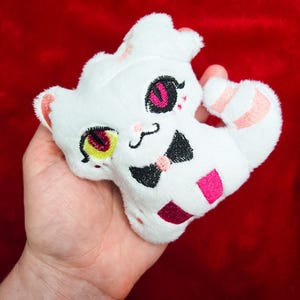 May include: A white plush toy in the shape of a cat, held in a hand. The cat has embroidered features including pink and black eyes, a black bow tie, and a pink and white striped tail. The background is a solid red.