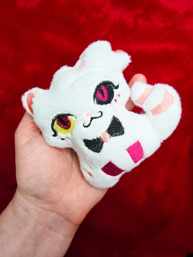 May include: A white plush toy in the shape of a cat, held in a hand. The cat has embroidered features including pink and black eyes, a black bow tie, and a pink and white striped tail. The background is a solid red.