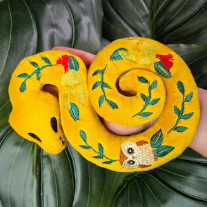 May include: A plush, yellow spiral-shaped toy with green embroidered vines and red flower accents. An embroidered owl is featured on the spiral. The toy is made of soft material.
