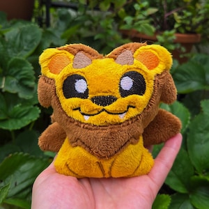 May include: A plush lion toy with a smiling face, featuring a yellow mane and body, brown accents, and black eyes. The toy is held in a hand against a backdrop of green foliage.