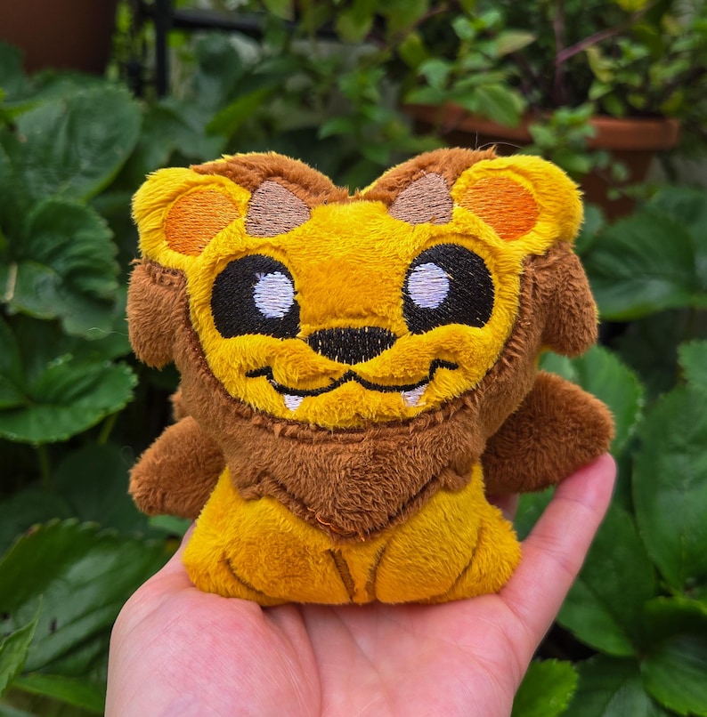 May include: A plush lion toy with a smiling face, featuring a yellow mane and body, brown accents, and black eyes. The toy is held in a hand against a backdrop of green foliage.