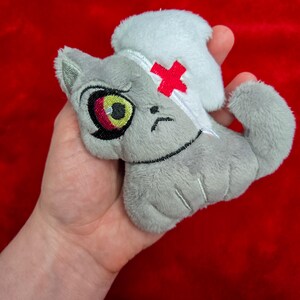 May include: A gray plush cat toy with a white nurse's cap and a red cross on its head. The cat has a sad expression with a large eye and a yellow iris. The toy is held against a red background.
