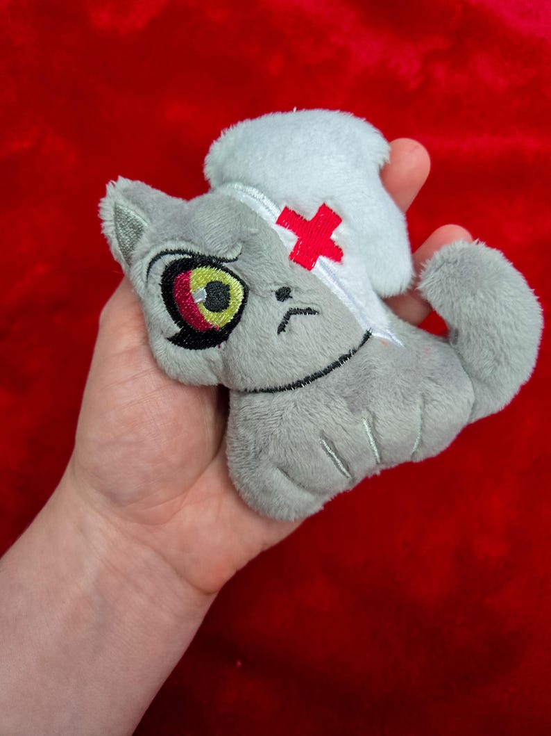 May include: A gray plush cat toy with a white nurse's cap and a red cross on its head. The cat has a sad expression with a large eye and a yellow iris. The toy is held against a red background.
