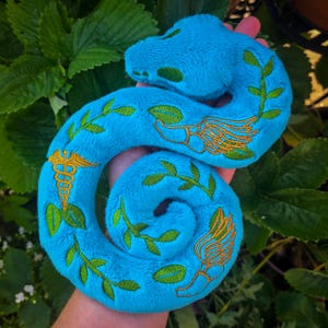 May include: A turquoise plush snake toy with green and gold embroidered details. The snake features leaf and wing designs, along with a medical symbol. The plush toy is held in a person's hand against a backdrop of green foliage.