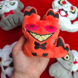 May include: A collection of plush cat toys in various colors. The central toy is orange with black accents, a stitched smile, and pink eyes. Other cats are white and gray, some with bowties and medical symbols.