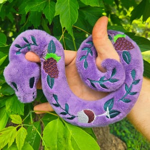 May include: A plush purple snake toy with embroidered green leaves and dark purple berry clusters. The snake's design includes a crescent moon and a wine glass. The toy is held in a hand against a backdrop of green leaves.