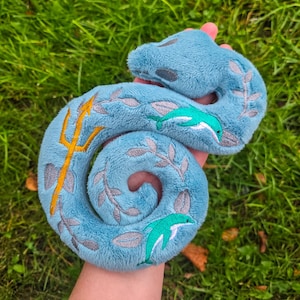 May include: A plush, light blue seahorse toy with embroidered details. The seahorse features a gold trident, gray leaf accents, and two green and white dolphins. The toy is held against a background of green grass.