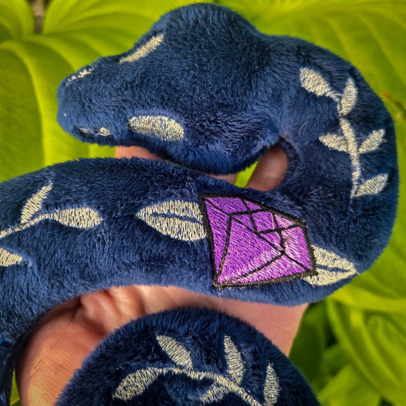 Op de afbeelding: Een marineblauwe pluche slangenspeelgoed met zilveren geborduurde bladeren en een paarse diamantvormige patch. De slang is zacht en knuffelig, perfect voor kinderen of verzamelaars.