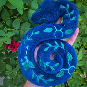 May include: A plush, coiled snake toy in a deep blue hue, adorned with embroidered teal-green leaves and celestial patterns. The snake's design includes a constellation and zodiac symbol. The soft toy is held against a backdrop of green foliage and a red rose.
