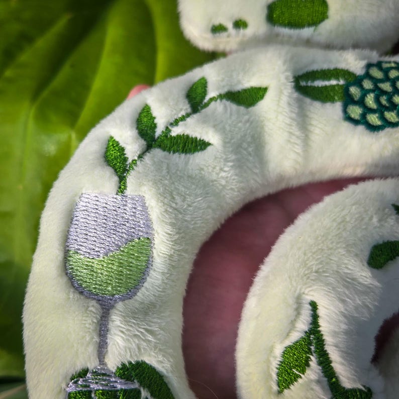 May include: A plush, off-white wine glass-shaped pillow with green and silver embroidered details. The pillow features a wine glass filled with green liquid, green leaves, and a cluster of grapes. The background is a blurred green.