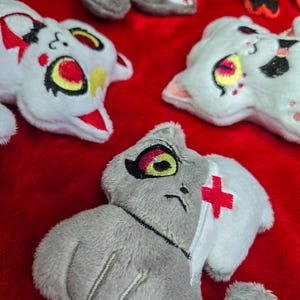 May include: A collection of plush cat toys in various colors, including white, gray, and orange, with embroidered details. The toys have large eyes and unique markings, set against a red background.