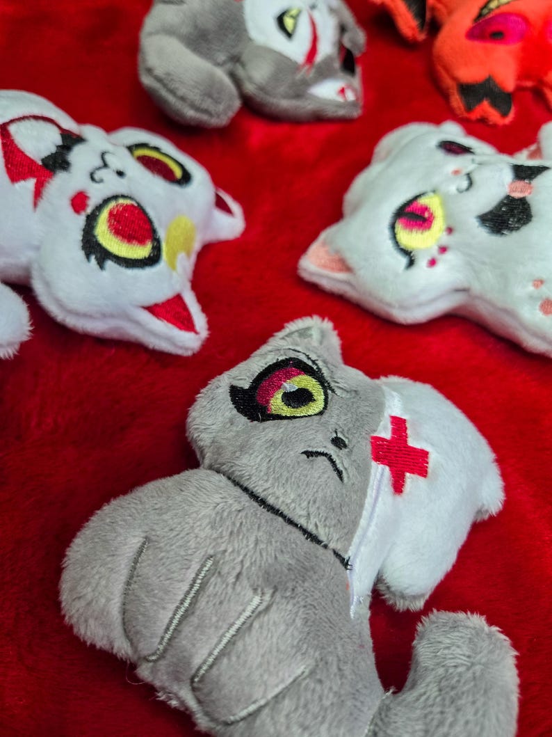 May include: A collection of plush cat toys in various colors, including white, gray, and orange, with embroidered details. The toys have large eyes and unique markings, set against a red background.