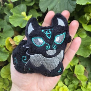 May include: A black plush cat with gray and teal embroidery. The cat has large eyes and a stylized design on its forehead. The plush toy is small and appears to be handmade.