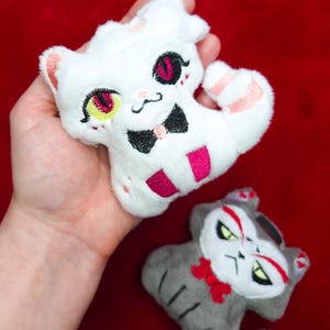 May include: Two plush cat toys. One is white with pink and black accents, a bow tie, and a striped tail. The other is gray with red and black accents and a red bow. The toys are on a red background.