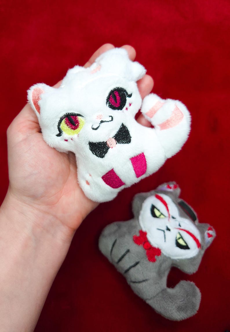 May include: Two plush cat toys. One is white with pink and black accents, a bow tie, and a striped tail. The other is gray with red and black accents and a red bow. The toys are on a red background.
