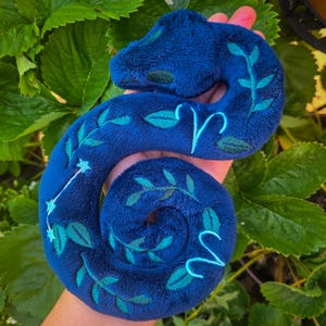 May include: A plush, dark blue Aries zodiac sign, featuring teal embroidered leaves and constellation details. The plush is held in a hand, with a background of green leaves.