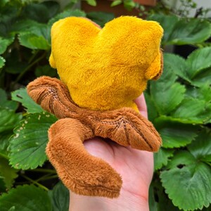 May include: A plush toy in the shape of a creature, featuring a yellow head and brown limbs. The toy is made of soft, textured fabric and is held in a hand against a backdrop of green leaves.