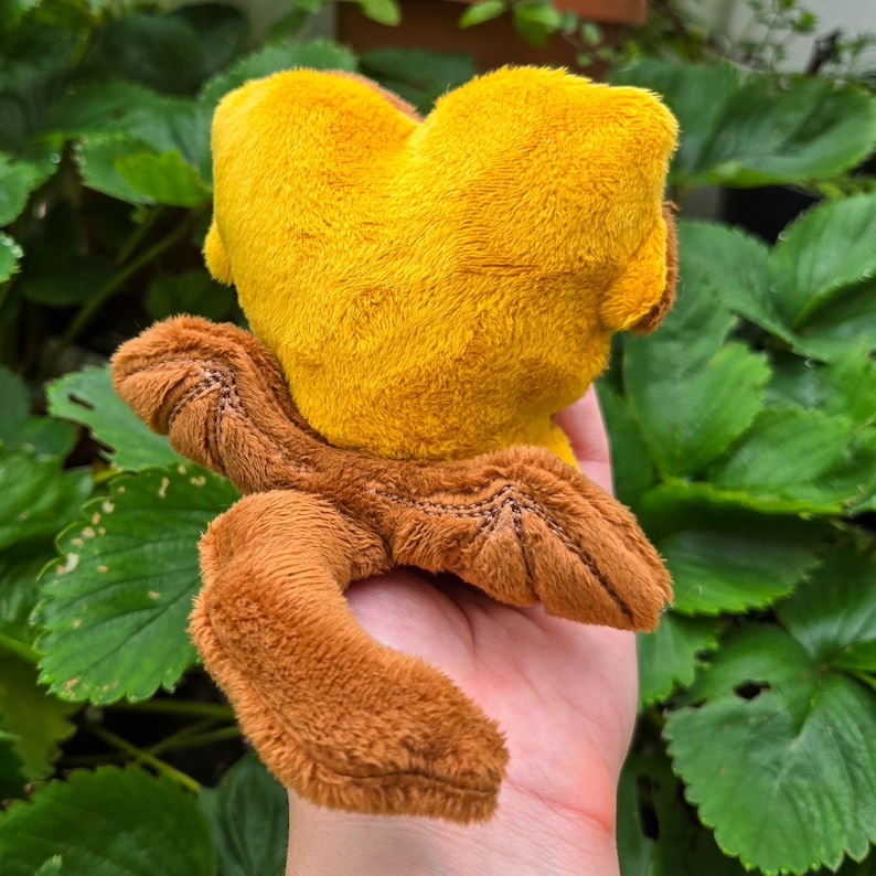 May include: A plush toy in the shape of a creature, featuring a yellow head and brown limbs. The toy is made of soft, textured fabric and is held in a hand against a backdrop of green leaves.