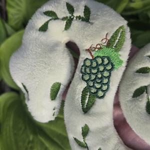 May include: A plush, white snake toy with green embroidered details. The snake features a green grape cluster and leaf design, with additional green leaf accents along its body. The toy is soft and cuddly, perfect for children.