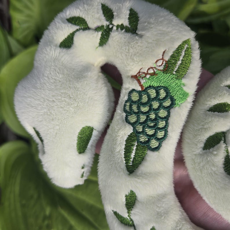 May include: A plush, white snake toy with green embroidered details. The snake features a green grape cluster and leaf design, with additional green leaf accents along its body. The toy is soft and cuddly, perfect for children.