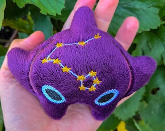 May include: A plush purple toy shaped like a manta ray, featuring embroidered yellow stars and a constellation design. The toy has two oval-shaped eyes with blue and black details.