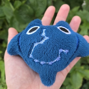 May include: A small, plush blue manta ray toy with embroidered details. The toy has black oval eyes and a stitched smile. The manta ray is held in a person's hand against a backdrop of green foliage.