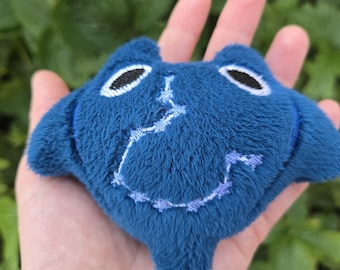 May include: A small, plush blue manta ray toy with embroidered details. The toy has black oval eyes and a stitched smile. The manta ray is held in a person's hand against a backdrop of green foliage.