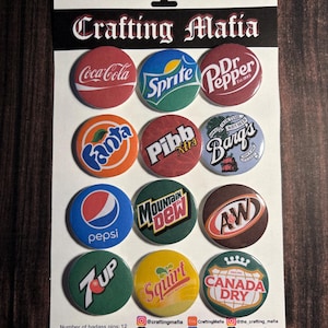 May include: Twelve 1.25 inch diameter round button pins featuring various vintage soda logos, including Coca-Cola, Sprite, Dr. Pepper, Fanta, Pibb Xtra, Barq's, Pepsi, Mountain Dew, A&W, 7 Up, Squirt, and Canada Dry. The pins are arranged in three rows of four on a white card with the text "Crafting Mafia" at the top.