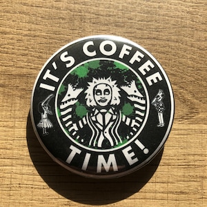 Peut inclure: Un bouton noir et vert avec le texte "It's Coffee Time!" et une image stylisée d'un monstre vert à trois têtes. Le monstre porte une chemise blanche et une cravate noire. Le bouton est entouré d'un cercle noir avec une bordure blanche.