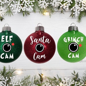Może przedstawiać: Trzy bombki choinkowe z brokatem, każda z innym motywem: elf, Święty Mikołaj i Grinch. Każda bombka ma białą ikonę aparatu i napis "Cam" pod spodem.