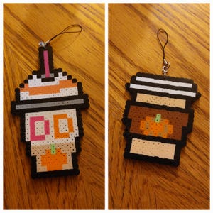 May include: Two pixel art charms: one resembling an iced coffee with orange, white, and pink details, and a pumpkin. The other charm is a coffee cup with a pumpkin design. Both charms have a black border and a string for hanging.