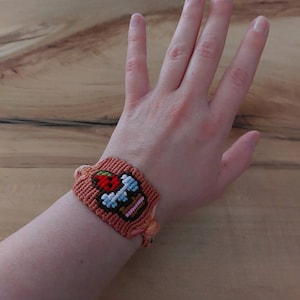 May include: A handmade macrame bracelet in shades of peach and brown, featuring a detailed embroidered design of a weightlifting barbell and a red apple. The bracelet is tied with peach-colored threads.