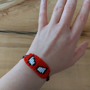May include: Red and black woven bracelet with white accents, worn on a wrist. The bracelet features a repeating pattern of two white rectangles inside black squares on a red background. The bracelet is handmade.