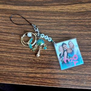 May include: A mermaid-themed charm with a gold-colored tail, a pearl, and a teal shell. The charm includes a small photo of four women and the text "H2O". The charm is attached to a black cord.