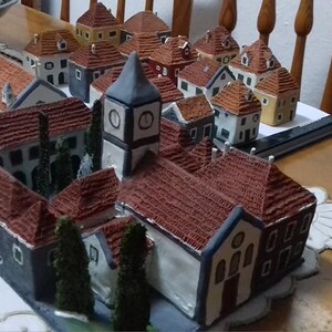 Może przedstawiać: Miniaturowa scena wiejska z r&oacute;żnymi małymi domkami, kościołem z wieżą zegarową i drzewami. Domki są wykonane z brązowej i białej ceramiki i mają czerwone dach&oacute;wki. Scena jest ustawiona na białym obrusie.