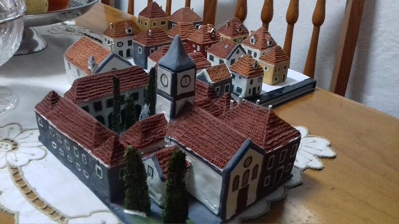 Może przedstawiać: Miniaturowa scena wiejska z r&oacute;żnymi małymi domkami, kościołem z wieżą zegarową i drzewami. Domki są wykonane z brązowej i białej ceramiki i mają czerwone dach&oacute;wki. Scena jest ustawiona na białym obrusie.