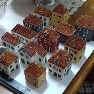 Może przedstawiać: Kolekcja ceramicznych domk&oacute;w w miniaturze, pomalowanych w r&oacute;żnych kolorach, w tym czerwonym, niebieskim, zielonym i ż&oacute;łtym. Domki mają szczeg&oacute;łowe dachy i okna, a niekt&oacute;re mają kominy. Domki są ułożone na białej powierzchni.