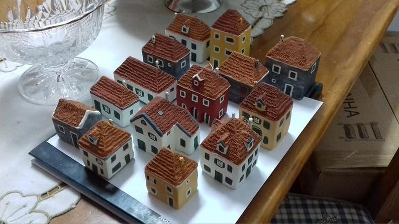 Może przedstawiać: Kolekcja ceramicznych domk&oacute;w w miniaturze, pomalowanych w r&oacute;żnych kolorach, w tym czerwonym, niebieskim, zielonym i ż&oacute;łtym. Domki mają szczeg&oacute;łowe dachy i okna, a niekt&oacute;re mają kominy. Domki są ułożone na białej powierzchni.
