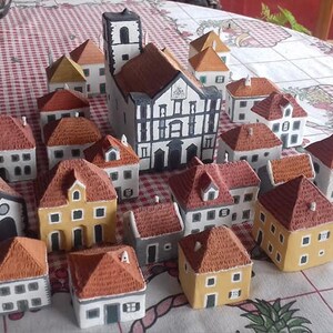 Może przedstawiać: Kolekcja miniaturowych domk&oacute;w ceramicznych w r&oacute;żnych stylach i kolorach, w tym duży kości&oacute;ł z biało-czarną fasadą. Domki są ułożone na szachownicy.