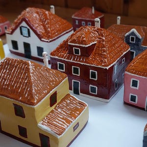 Może przedstawiać: Kolekcja miniaturowych domk&oacute;w ceramicznych w r&oacute;żnych kolorach, w tym czerwonym, ż&oacute;łtym, r&oacute;żowym i brązowym. Domki mają szczeg&oacute;łowe dachy i okna, a niekt&oacute;re mają kominy. Domki są ułożone na białej powierzchni.