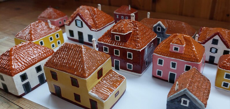 Może przedstawiać: Kolekcja miniaturowych domk&oacute;w ceramicznych w r&oacute;żnych kolorach, w tym czerwonym, ż&oacute;łtym, r&oacute;żowym i brązowym. Domki mają szczeg&oacute;łowe dachy i okna, a niekt&oacute;re mają kominy. Domki są ułożone na białej powierzchni.